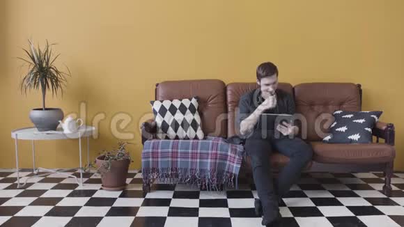 英俊的休闲年轻人在家里使用平板电脑坐在客厅的沙发上浏览互联网股票视频的预览图