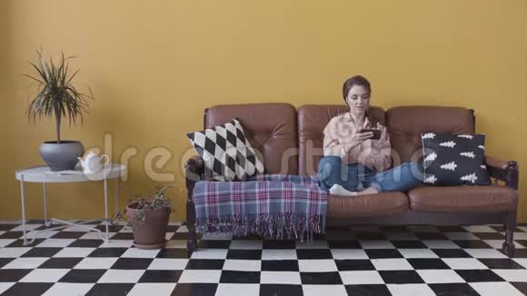 快乐的年轻美丽的女人在她的智能手机上玩游戏坐在明亮时尚的房间里的沙发上视频的预览图