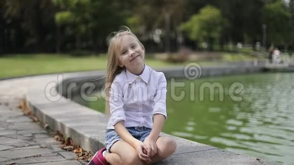 可爱的金发女孩坐在湖岸上把头发从她的脸上移开骑着滑板车视频的预览图