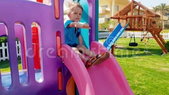 在公园儿童游乐场里一个快乐微笑的幼儿男孩骑在滑梯上视频的预览图