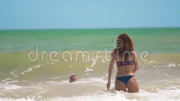 夏天阳光明媚的一天这个快乐的晒黑孩子在热带海滩和妈妈一起在海浪中快乐地游泳和跳跃视频的预览图