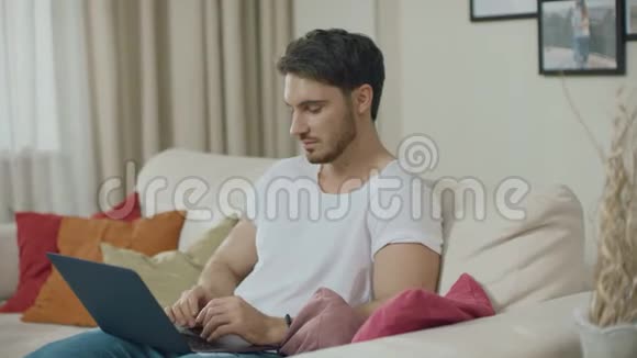 微笑的男人在家里的沙发上用笔记本电脑工作年轻人在网上聊天视频的预览图