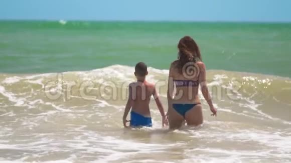 夏天阳光明媚的一天这个快乐的晒黑孩子在热带海滩和妈妈一起在海浪中快乐地游泳和跳跃视频的预览图