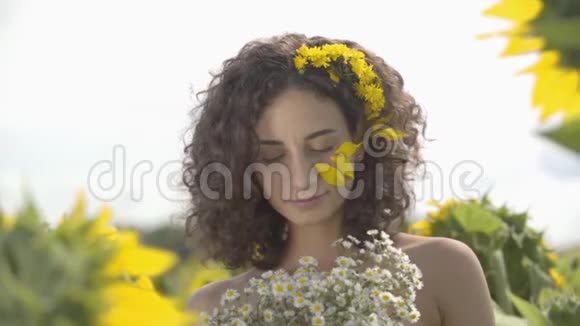 一个美丽的卷曲女孩的肖像看着相机抚摸着她的头发站在向日葵的田野上带着她的头发视频的预览图