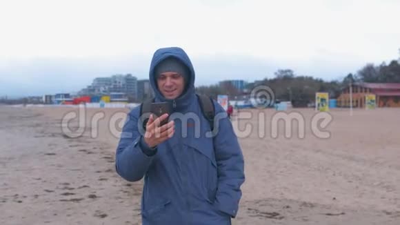 男人博主穿着蓝色羽绒服在海边沙滩上散步在手机上聊天视频的预览图