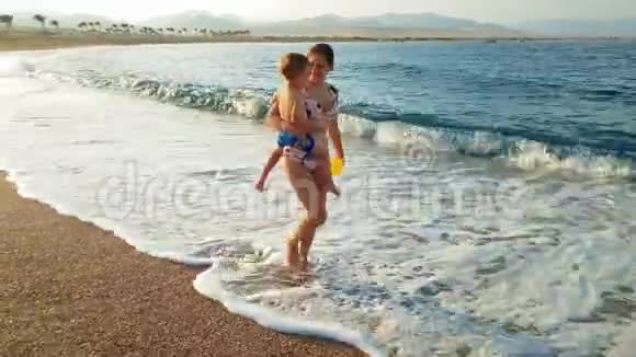 4k视频快乐微笑的年轻女子抱着她的小儿子在日落时分在海滩上散步视频的预览图