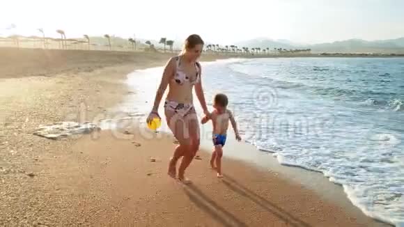 4k镜头日落时分快乐的牧羊妈妈带着蹒跚学步的儿子在沙滩上奔跑视频的预览图