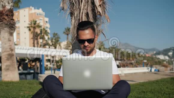 年轻的留胡子男子t恤戴着眼镜坐在公园的草地上手里拿着笔记本电脑写着旅游信息视频的预览图