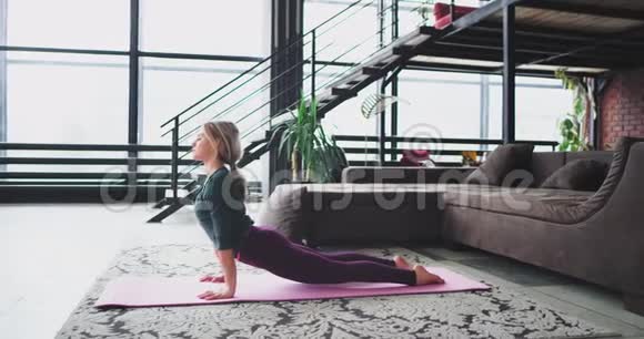在客厅阁楼的垫子上有魅力的女人在家里伸展身体练习瑜伽视频的预览图