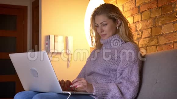 特写侧景拍摄的成年白种人迷人的女性在笔记本电脑上打字而坐在室内的沙发上视频的预览图