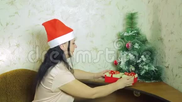 一个戴着圣诞老人帽子的微笑的女人正悄悄地把圣诞礼物放在圣诞树下视频的预览图