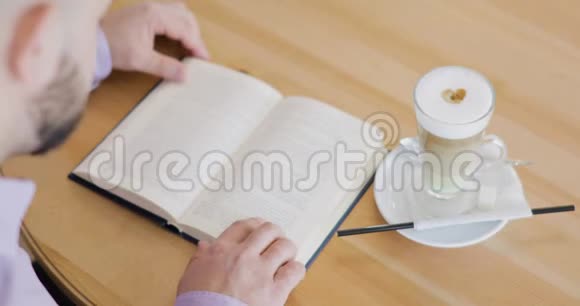 男人正在看一本书手指在书页上坐在咖啡屋里喝着一杯卡布奇诺快速阅读视频的预览图