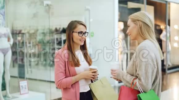 可爱的女孩朋友正在购物中心谈笑风生手里拿着外卖饮料和五颜六色的纸袋快乐视频的预览图