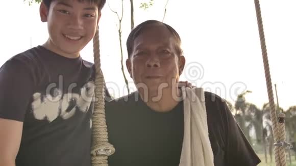 老人和年轻男孩微笑着的肖像4K手持亚洲男孩与大父亲在秋千上看镜头视频的预览图
