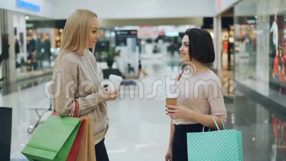 美丽的年轻女性在购物中心聊天手里拿着外卖饮料和纸袋讨论购买和购买视频的预览图