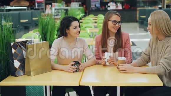 可爱的女孩坐在购物中心的咖啡馆里拿着饮料和纸袋谈论分享新闻妇女视频的预览图