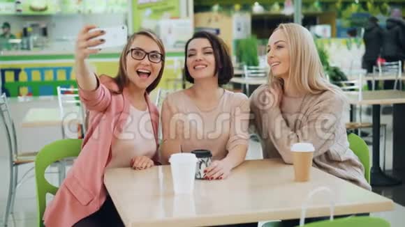 漂亮的年轻女士正在使用智能手机自拍坐在咖啡馆的桌子上用饮料摆姿势友谊视频的预览图