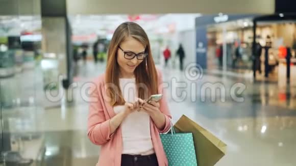 美丽的头发可爱的女孩正在使用智能手机和微笑而步行在购物中心与纸袋互联网视频的预览图