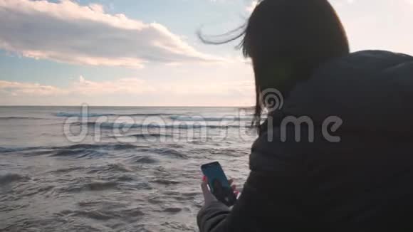 年轻的黑发女人坐在岩石海滩上打电话自拍视频的预览图