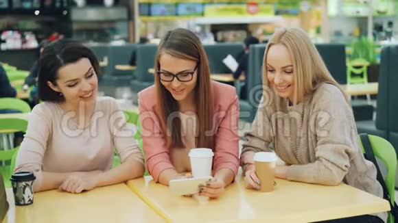 快乐的年轻女士们一起使用智能手机看着屏幕微笑着笑着坐在咖啡馆购物视频的预览图