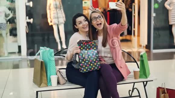 可爱的女孩正在购物中心自拍摆着纸袋时髦的手势坐在长凳上视频的预览图