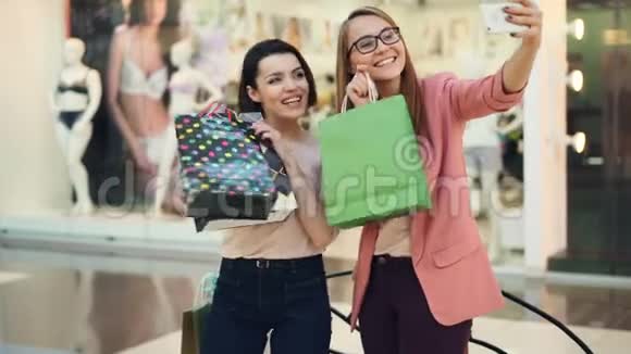 快乐的购物狂们在购物中心用纸袋自拍使用智能手机相机手持设备和摆姿势视频的预览图