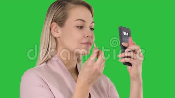 一位美丽的金发女郎她正在用红色口红涂嘴唇在绿色的屏幕上看着手机视频的预览图