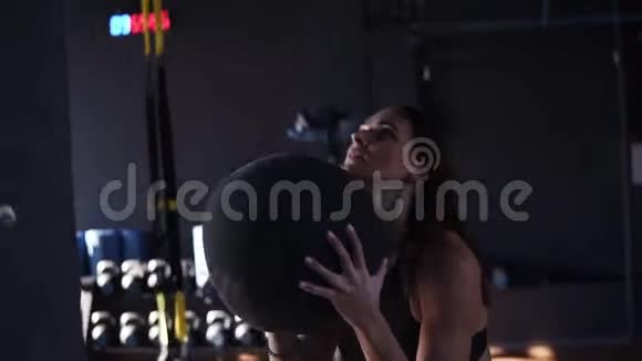 活跃的年轻的黑发女人蹲在一个深色的现代健身房里把一个药球扔到墙上视频的预览图