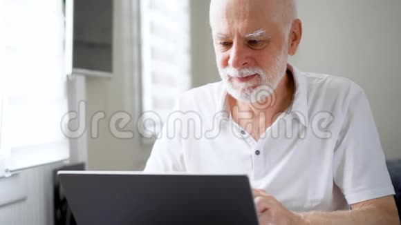 英俊的老人在家里用笔记本电脑工作收到好消息兴奋和高兴视频的预览图