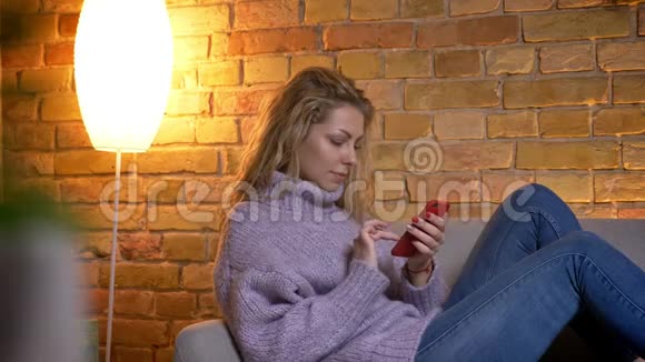 成年白种人金发女性在沙发上悠闲地坐在室内休息时浏览电话的特写镜头视频的预览图