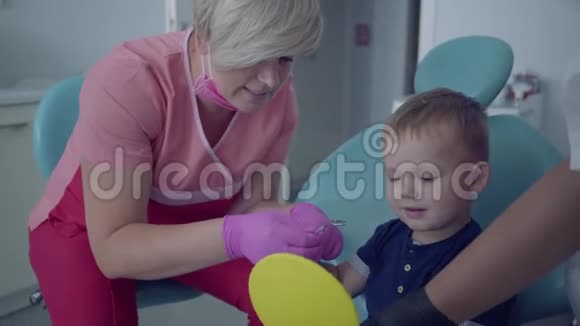 戴着医用口罩和手套的牙医准备检查椅子上坐着镜子的无忧无虑的小男孩的牙齿视频的预览图