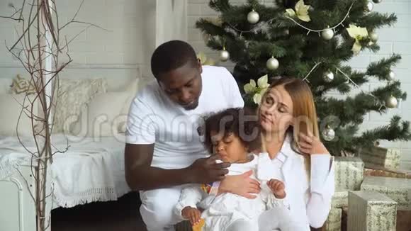 年轻幸福的多民族家庭母亲父亲和穿着白色衣服的小女儿坐在圣诞树旁视频的预览图