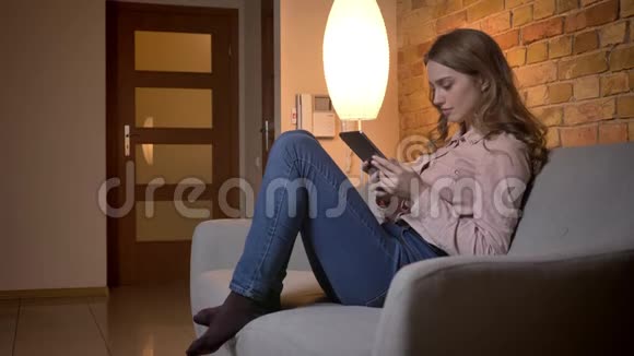 年轻的金发少女坐在沙发上在舒适的家中专心地看着平板电脑的画像视频的预览图