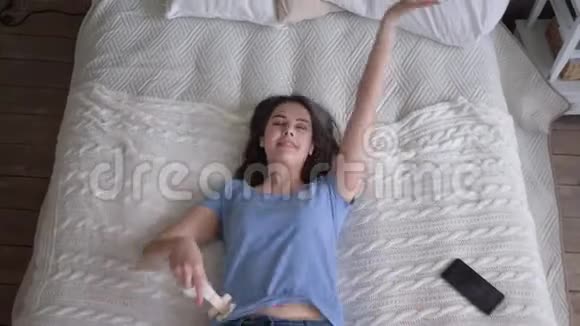 戴着耳机的快乐女孩躺在床上听着智能手机上的歌曲在家放松时欣赏音乐视频的预览图