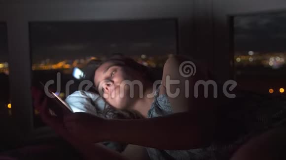 无聊的年轻漂亮女人躺在床上在黑暗中滚动他的手机电话里的灯是视频的预览图