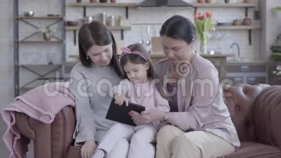 祖母母亲和小女儿一起坐在现代工作室公寓的沙发上那个拿着小玩意的女孩视频的预览图