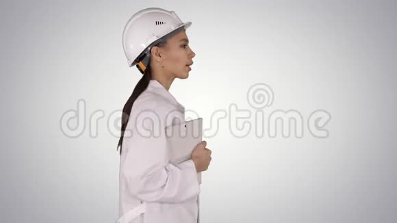 穿着白色实验室外套和戴着白色安全安全帽的西班牙裔有魅力的女士她穿着渐变的笔记本或平板电脑走路视频的预览图