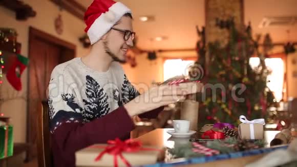 家中的杉树旁一个留着胡子的人坐在礼物上打个蝴蝶结戴着圣诞帽的家伙视频的预览图