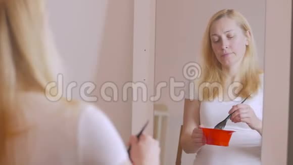 一位金发女人坐在镜子前的红色塑料碗里正在准备一种混合物来减轻头发的颜色视频的预览图