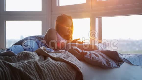 躺在床上的年轻美女在令人惊叹的日落中使用手机并具有镜头耀斑效果慢慢慢慢视频的预览图