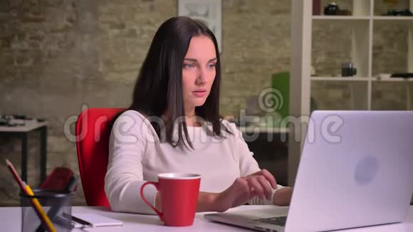 女商人在笔记本电脑前的办公室工作喝咖啡同时观看屏幕视频的预览图