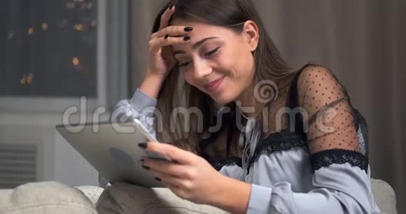 有着数码平板电脑的漂亮女人坐在家里的沙发上视频的预览图