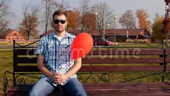 戴着黑色眼镜的野蛮年轻人坐在一张带着红色气球的长凳上看着相机复制空间慢动作视频的预览图