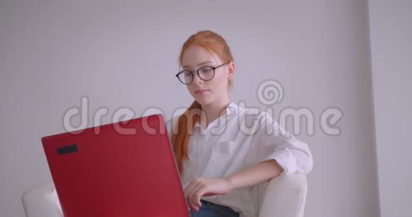 戴着眼镜的年轻漂亮的白种人红发女商人用笔记本电脑坐在扶手椅上的特写照片视频的预览图