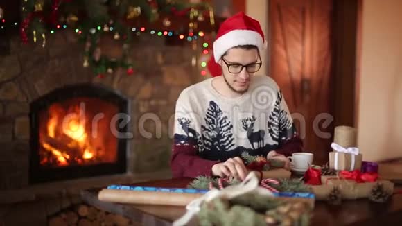 大胡子坐着在壁炉旁的新年礼物上系一个蝴蝶结看着相机穿着圣诞衣服视频的预览图