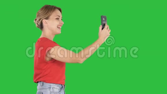 美丽的年轻女子走着拿着智能手机在绿色屏幕上拍照和自拍色度键视频的预览图