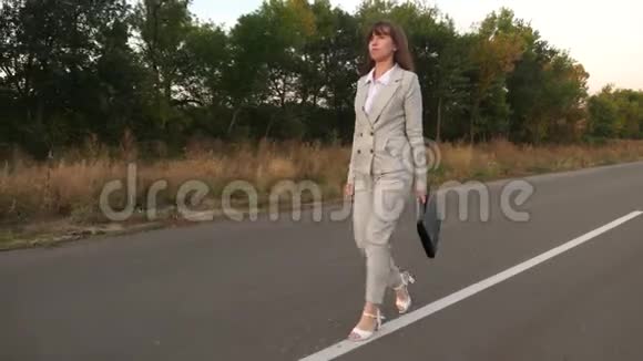 女商人带着黑色公文包穿着浅色西装穿着白色高跟鞋沿着柏油路走到城外视频的预览图