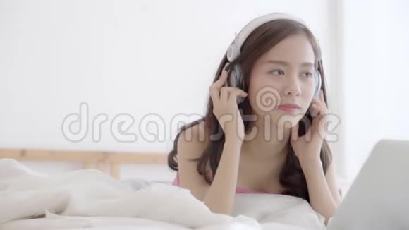 美丽的年轻亚洲女人躺在卧室里用手提电脑放松听音乐女孩在家里看视频聊天视频的预览图