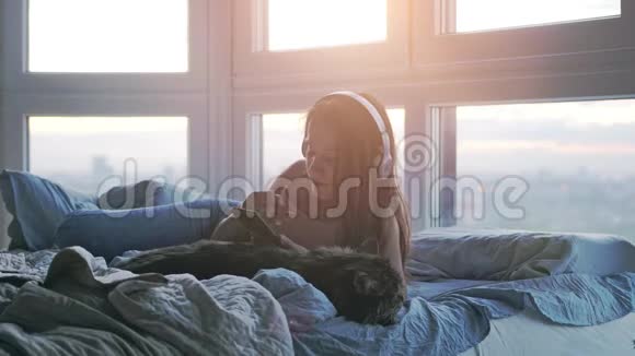 年轻的黑发女子戴着耳机与她可爱的缅因州考恩猫放松听音乐躺在床上使用移动视频的预览图