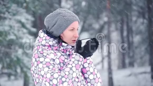 一个年轻的旅游女孩正在用热茶咖啡温暖自己站在一个下雪的冬天的森林或公园里妇女视频的预览图